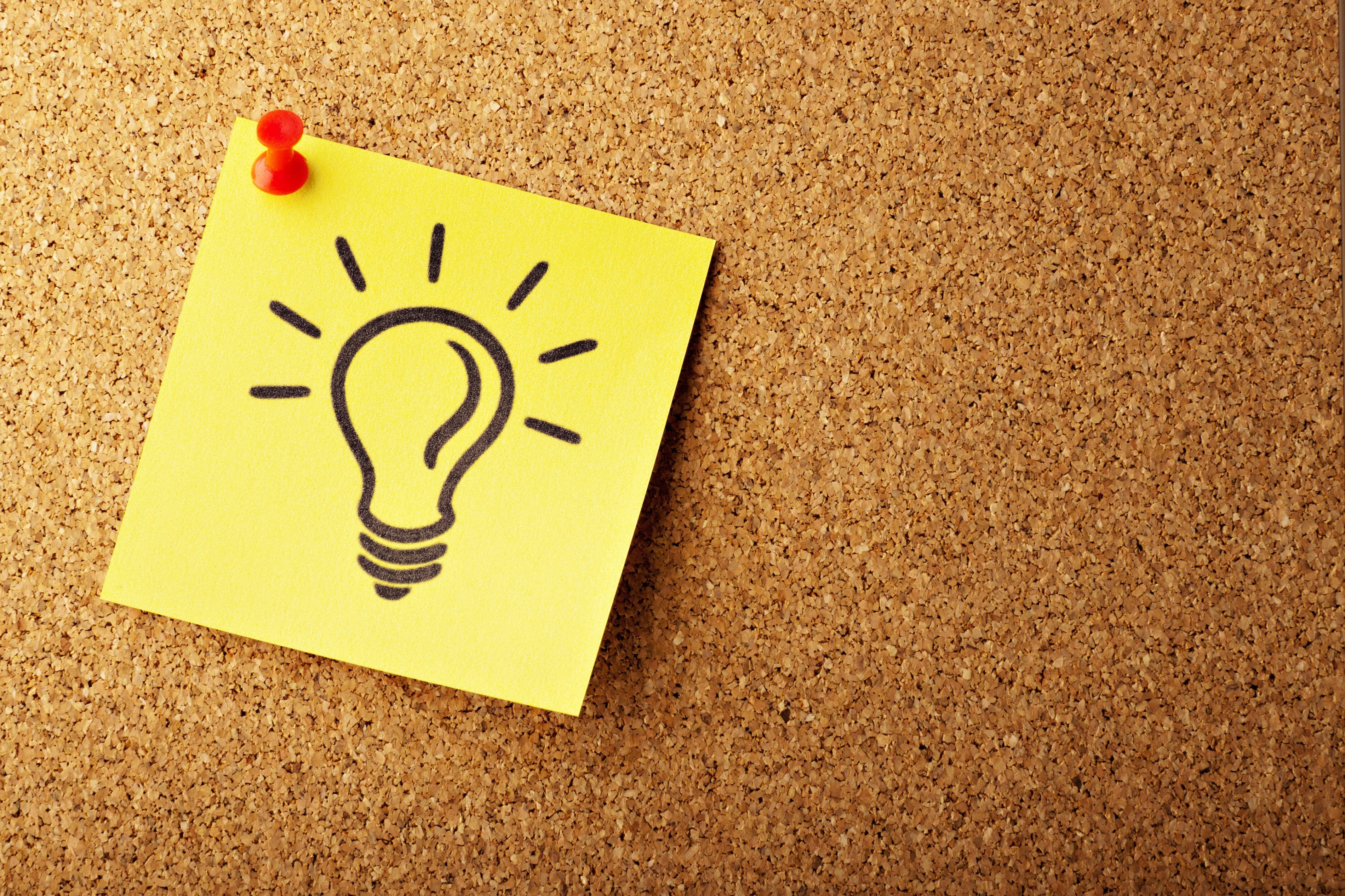 A yellow sticky note with a lightbulb drawing is stuck to a corkboard.