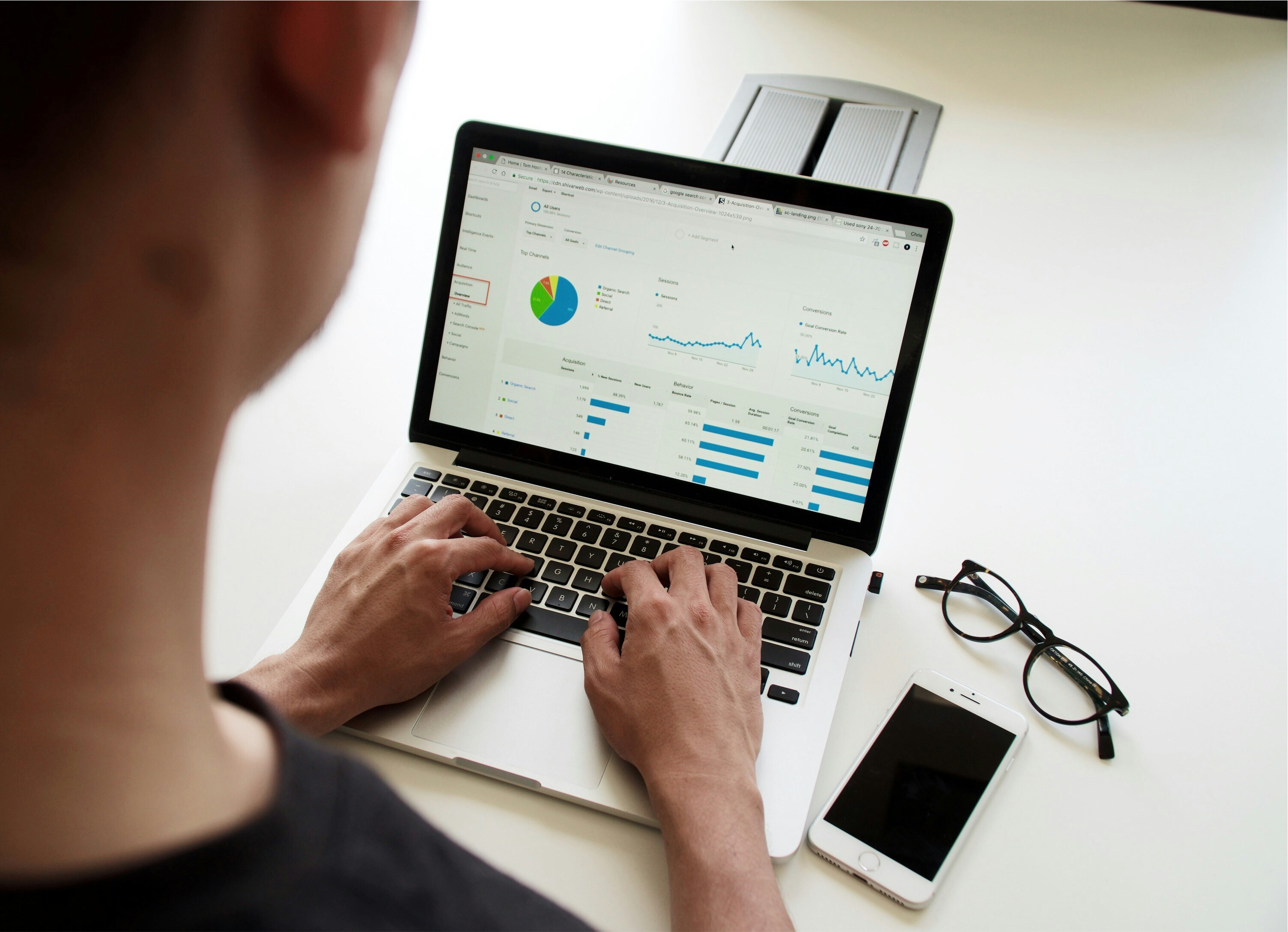 A person is typing on a laptop. There is data pulled up on the screen while a phone and glasses are set on the table.