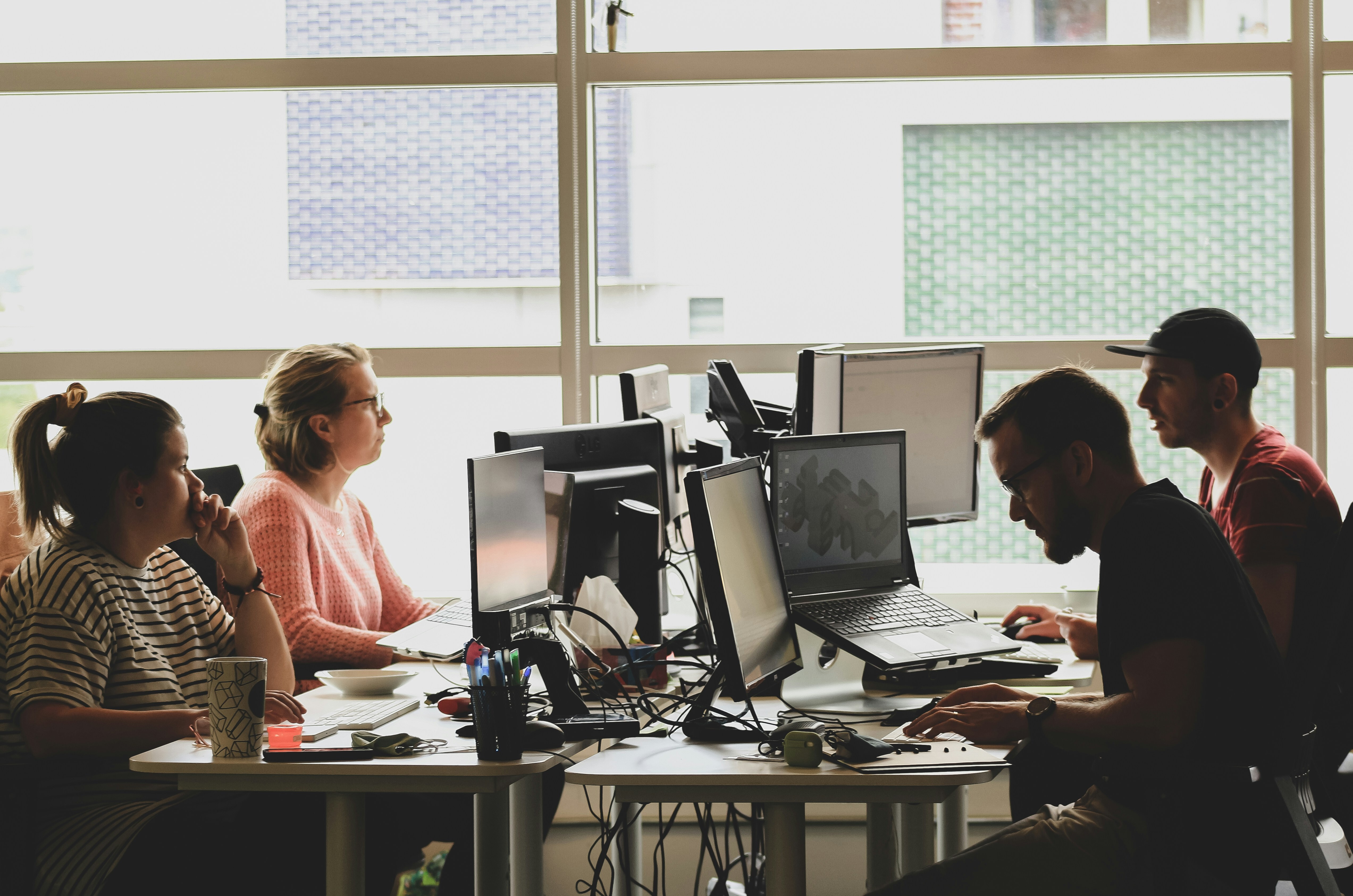 A group of four people are sitting at desks in an office environment. They are all looking at their computers.