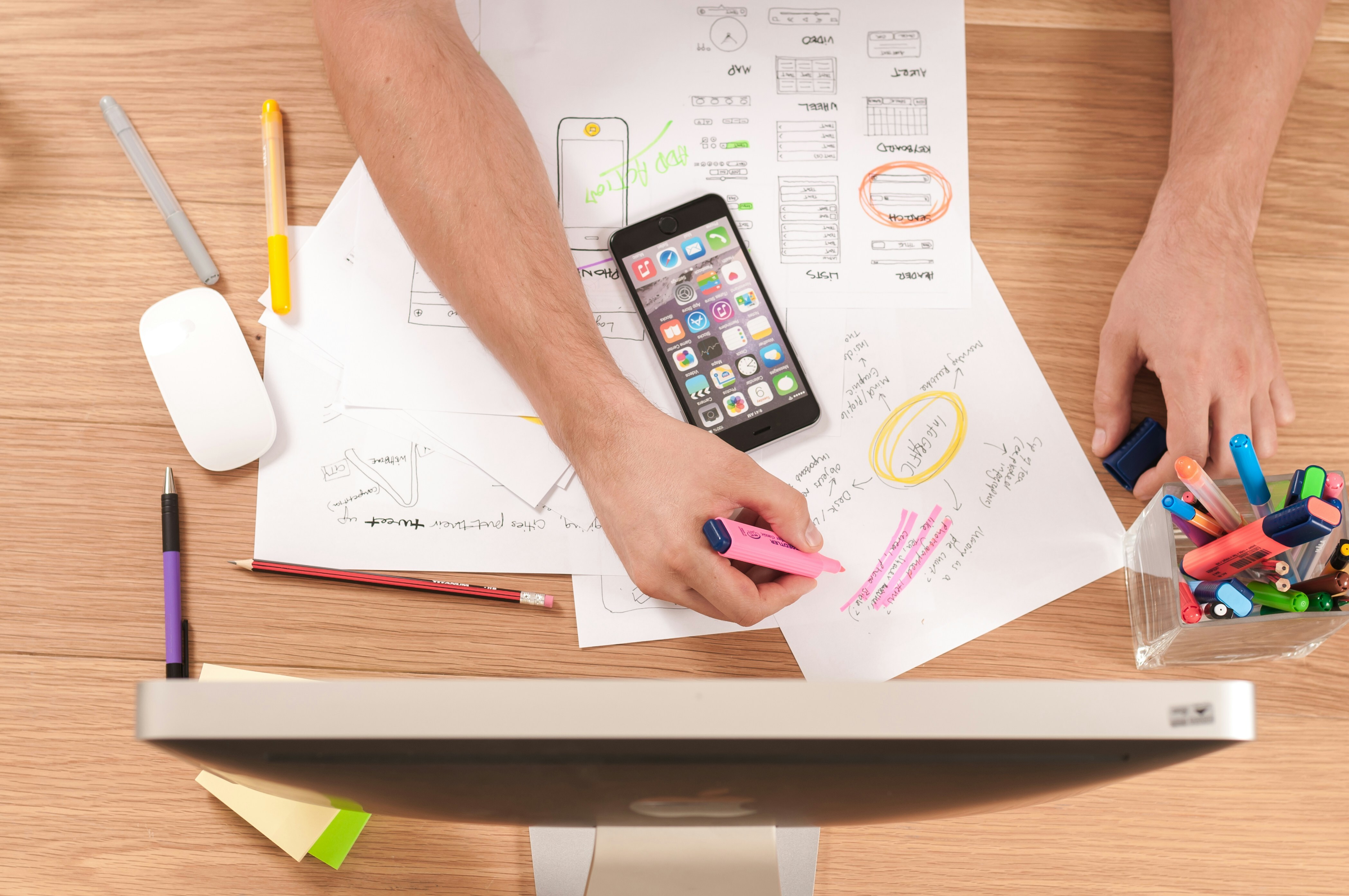 A desk has a computer, papers, pens, markers and pencils on it. A person is highlighting words in pink marker. There is a phone placed on the papers.