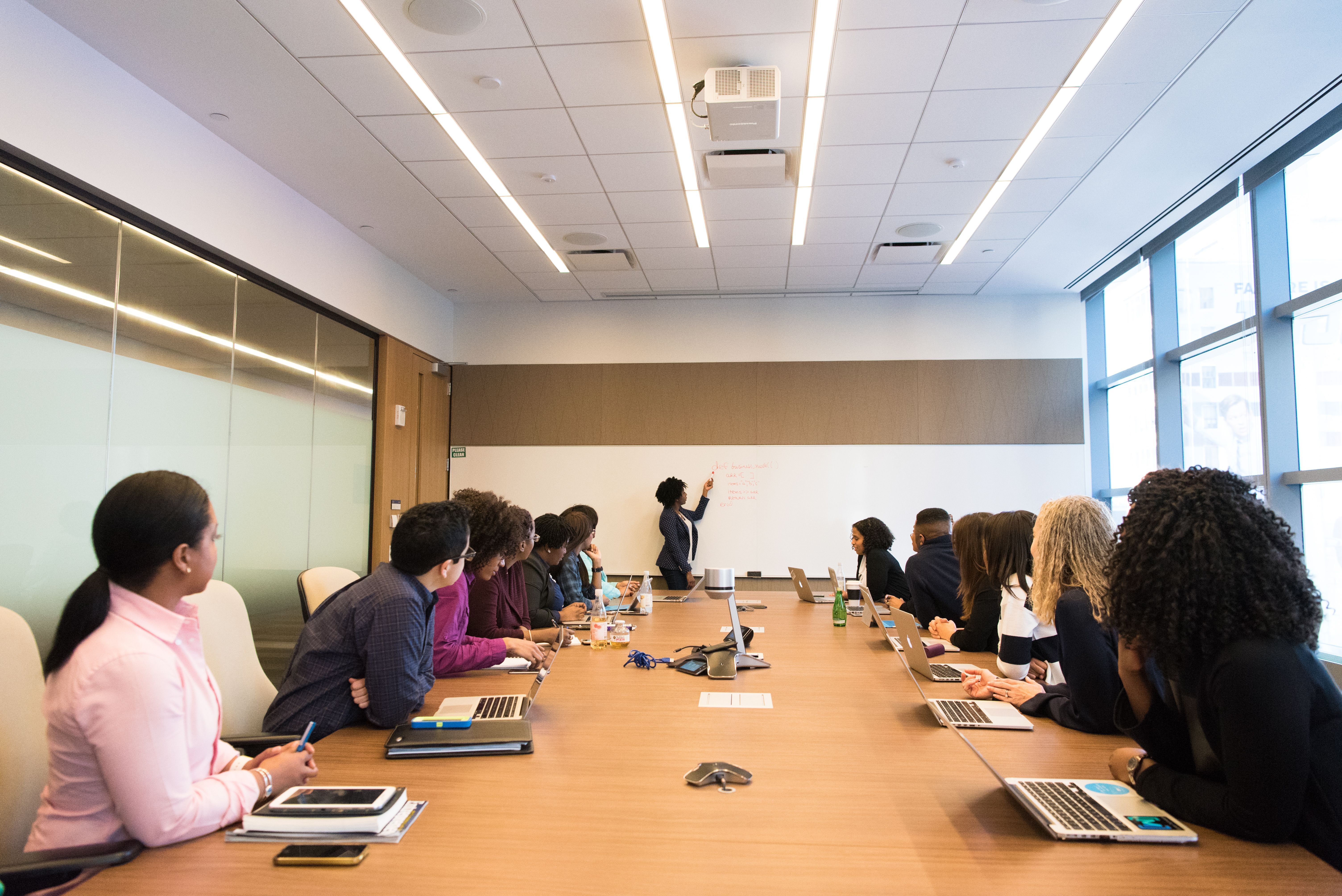 A large group of people are gathered around a large conference table, watching a presenter write on a whiteboard.