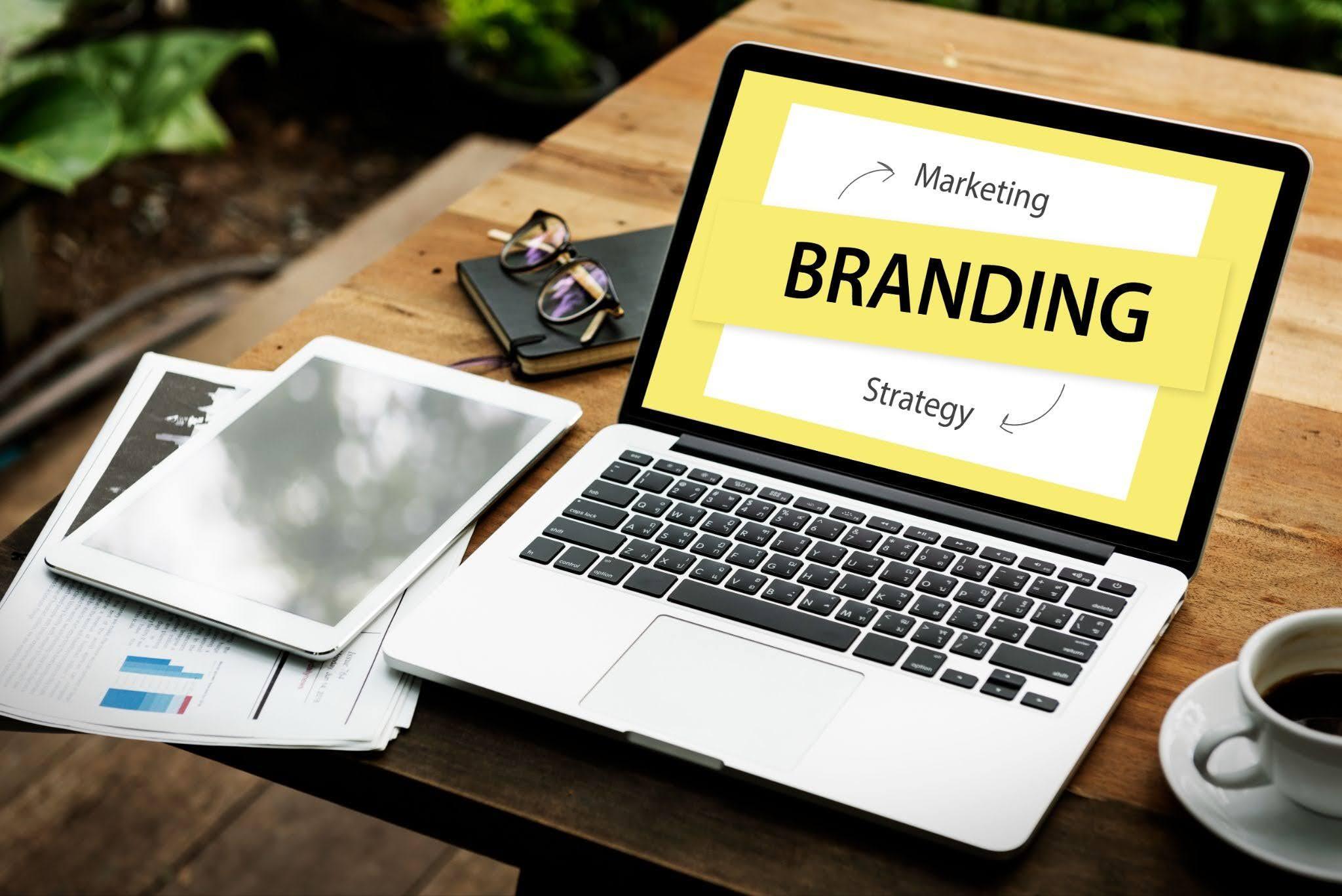 A laptop screen reads “Marketing Branding Strategy” in black font against a white and yellow background. A tablet, coffee mug, notebook, glasses and sheets of paper are placed on the table.